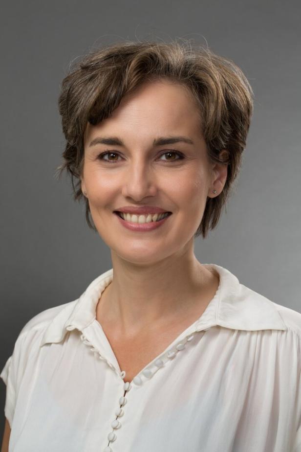 Eine lächelnde Frau mit kurzem, braunem Haar und einem weißen Hemd vor einem grauen Hintergrund.