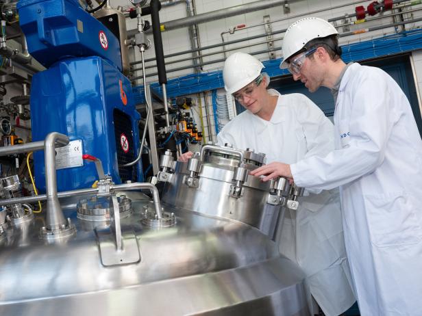Zwei Personen in Laborkitteln und Schutzhelmen arbeiten an einem großen, silbernen Tank.