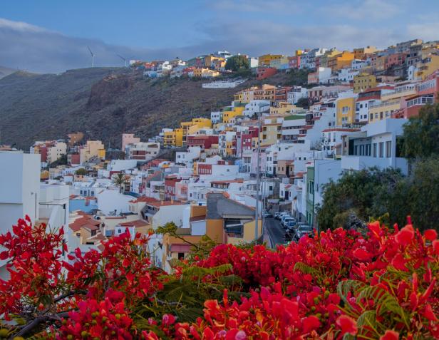 Blick auf San Sebastián de La Gomera mit bunten Häusern und roten Blüten im Vordergrund.
