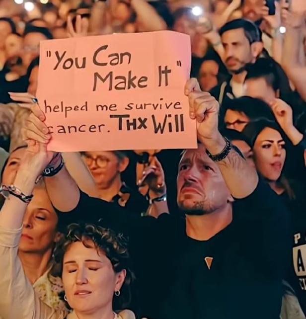 Ein Mensch hält ein Schild mit der Aufschrift „You Can Make It helped me survive cancer. Thx Will“ in die Höhe.