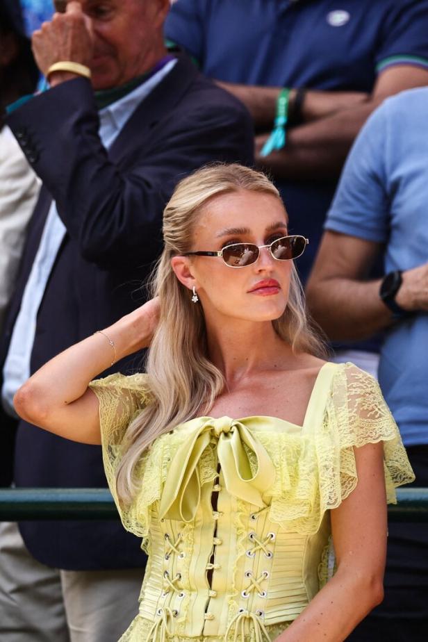 Eine Frau mit Sonnenbrille und gelbem Kleid sitzt im Publikum eines Tennisturniers in Wimbledon.
