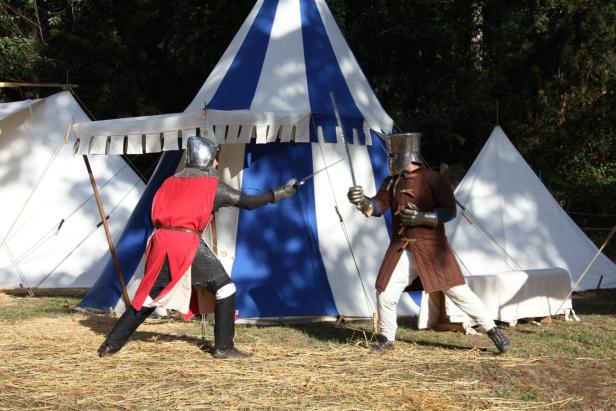 Zwei Männer in altertümlichen Gewändern kämpfen mit Schwertern vor einem Zelt.