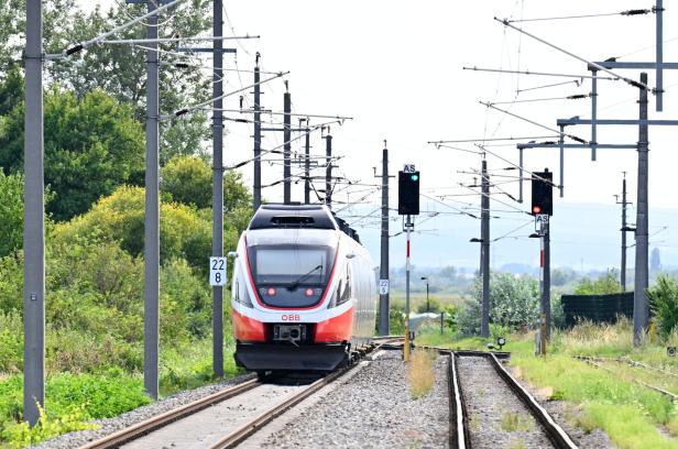 ÖBB-Zug fährt auf elektrifizierter Strecke mit Oberleitungen und Signalen durch eine grüne Landschaft.