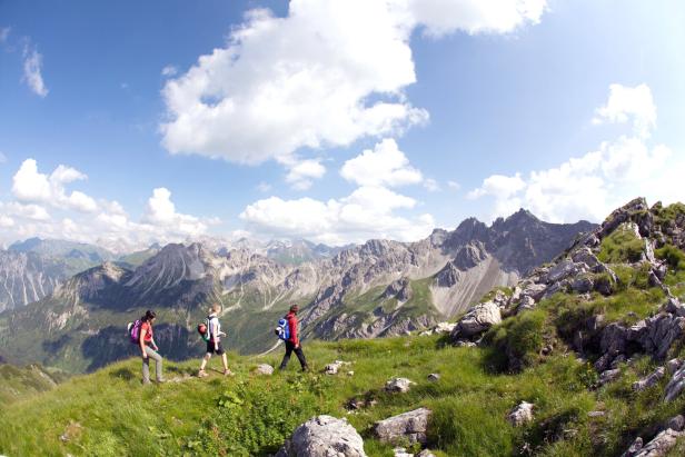 Drei Wanderer gehen auf einem grasbewachsenen Bergrücken mit Blick auf eine Bergkette.