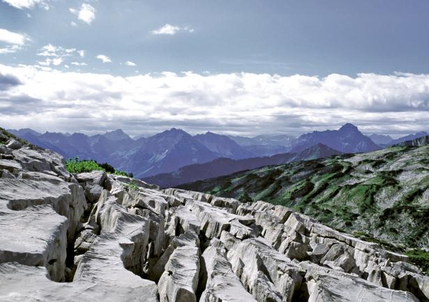 Blick über schroffe Felsen auf eine Berglandschaft unter bewölktem Himmel.