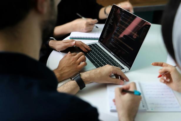 Mehrere Personen sitzen rund um einen Laptop an einem Tisch und schreiben Notizen auf Zetteln.