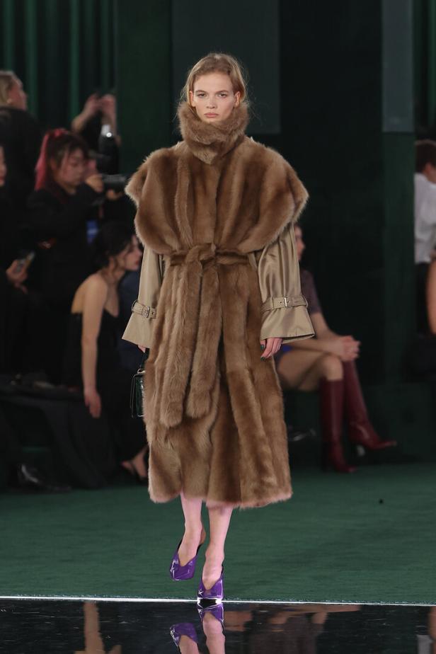 Ein Model präsentiert einen braunen Pelzmantel und violette Schuhe auf dem Laufsteg.