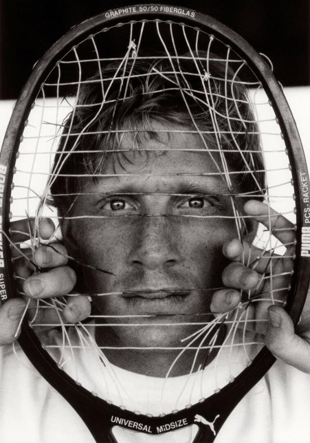 Ein Schwarzweißporträt von Boris Becker, der einen Tennisschläger mit gerissenen Saiten vor sein Gesicht hält.