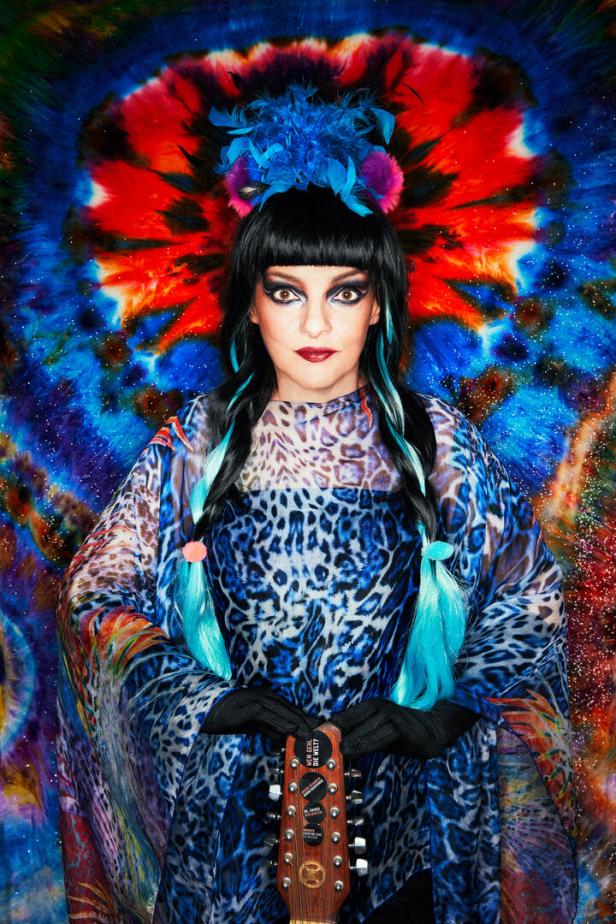 Eine Frau mit blau gefärbten Zöpfen hält eine Gitarre vor einem psychedelischen Hintergrund.