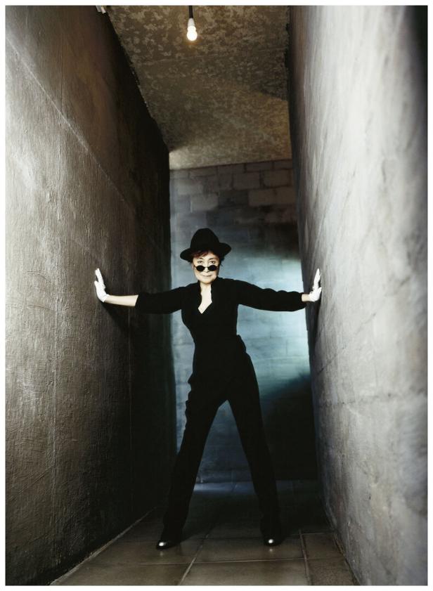 Yoko Ono posiert in einem schwarzen Anzug und Hut in einem engen Gang.