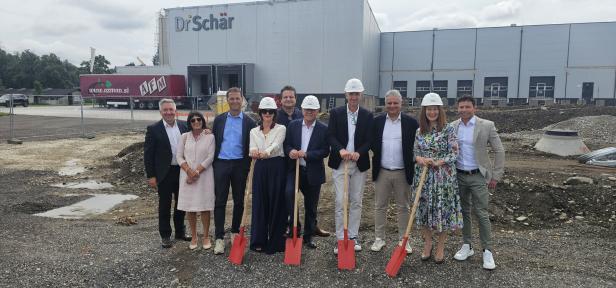 Der Dr. Schär Standort Klagenfurt wird ausgebaut. Menschen mit Spaten und Bautellenhelmen signalisieren die kommende Errichtung einer neuen Lagerhalle.