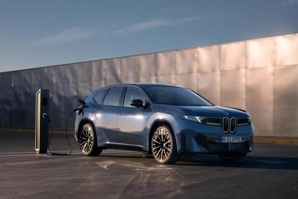 Der neue blaue BMW iX3, seitlich von der Sonne beleuchtet, neben einer Chromwand und einer E-Ladestation.
