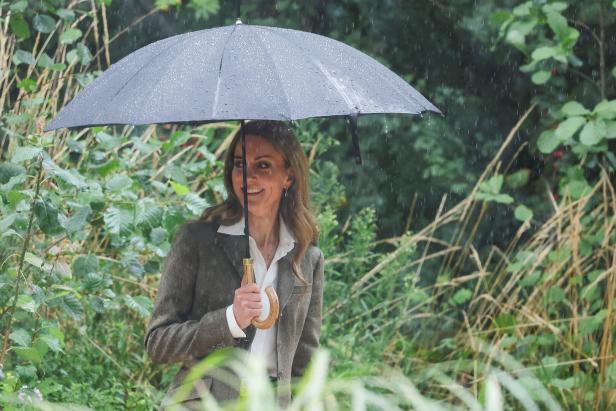 Zwischenzeitlich kam Kate in den Regen Prinzessin Kate lächelt im Regen unter einem schwarzen Regenschirm, umgeben von Grün.