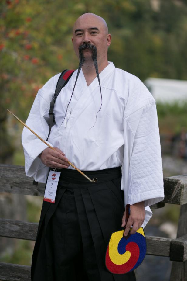 Ein Mann mit langem, dünnem Schnurrbart in traditioneller japanischer Kleidung hält einen Fächer und eine Pfeife.