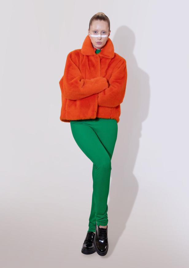 Eine Frau mit weißer Farbe im Gesicht trägt eine orangefarbene Jacke und grüne Leggings.