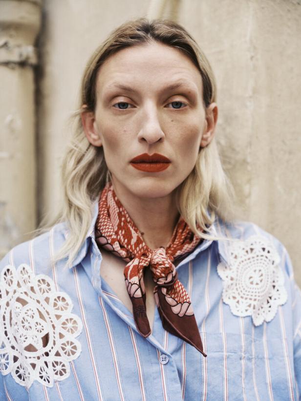 Eine Frau mit blonden Haaren und rotem Lippenstift trägt ein blau gestreiftes Hemd und ein Halstuch.