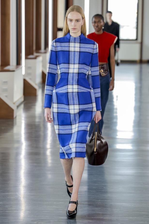 Ein Model läuft in einem blau-weiß karierten Kleid und mit einer braunen Handtasche über einen Laufsteg.