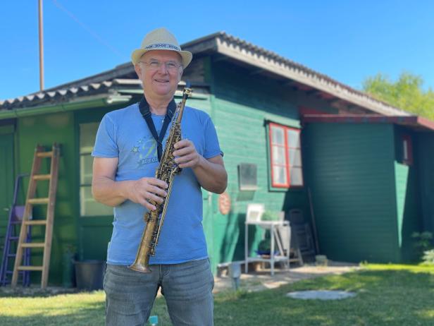Ein Mann mit Hut und Saxophon steht vor einem grünen Haus im Garten.