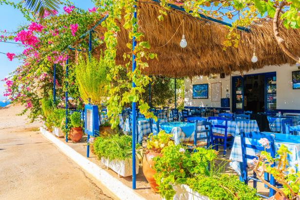 Einladendes griechisches Restaurant mit blau-weißen Tischen und üppiger Blumenpracht.