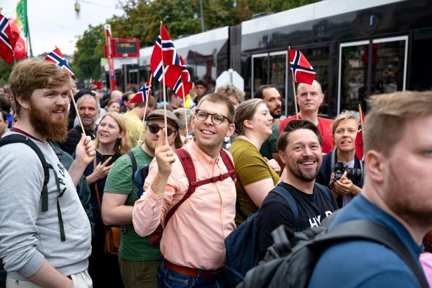 Eine fröhliche Menschenmenge hält norwegische Flaggen und steht vor einer Straßenbahn.