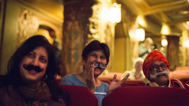 Drei Personen sitzen im Theater und halten Schnurrbärte vor ihre Gesichter.