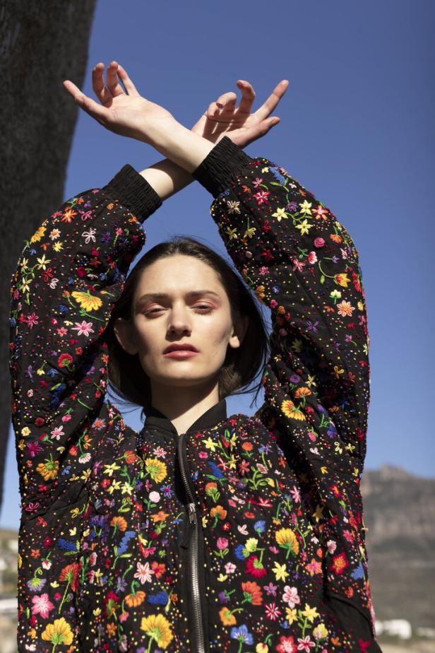 Eine Frau mit erhobenen Händen trägt eine schwarze Jacke mit bunten Blumenstickereien.