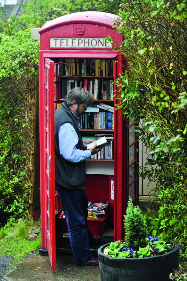Ein Mann steht in einer Telefonzeller, die voll mit Büchern ist, und liest.