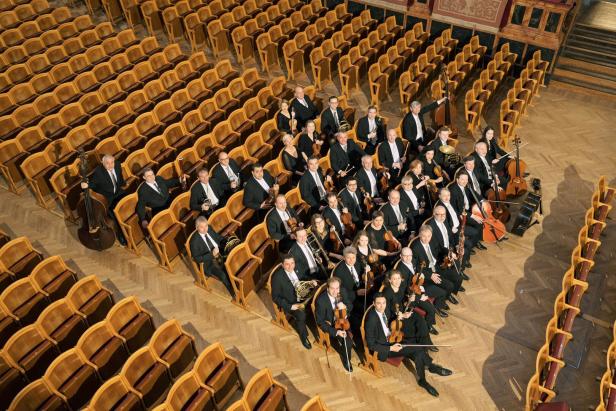 Ein Orchester sitzt in einem Konzertsaal zwischen den Publikumsreihen.