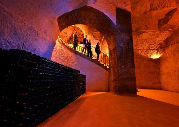 Menschen steigen eine Stufe in einen riesigen Keller hinab. Unten stehen Regale voll mit Champagnerflaschen.