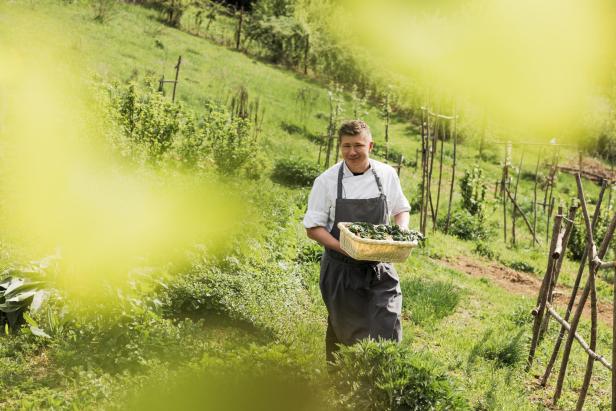 Ein Koch trägt einen Korb mit frischem Gemüse auf einem grünen Feld.