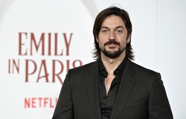 Lucas Bravo mit Bart und dunklem Anzug steht vor dem Schriftzug "Emily in Paris" und Netflix.