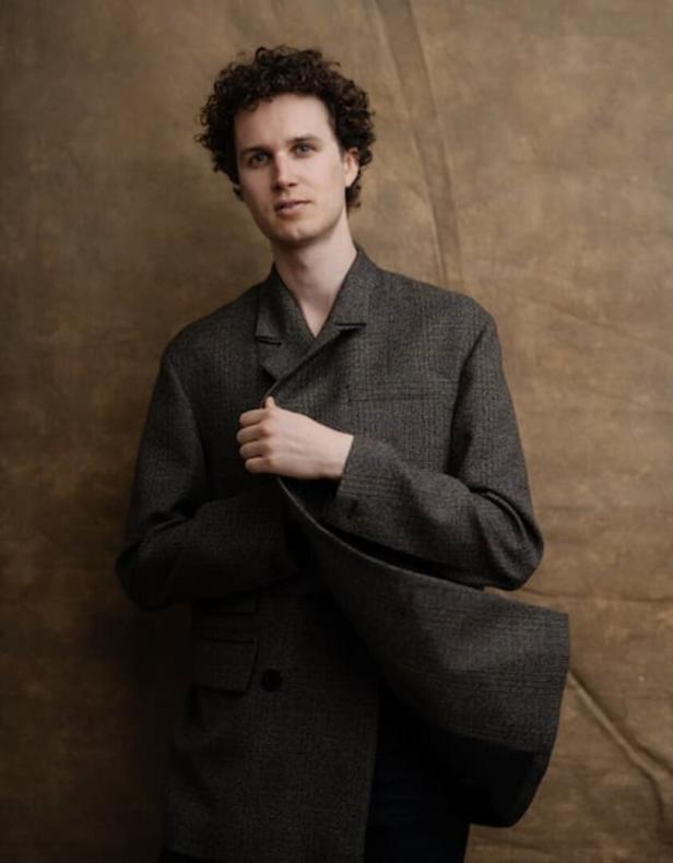 Ein Mann mit lockigem Haar trägt einen übergroßen Blazer vor einem braunen Hintergrund.