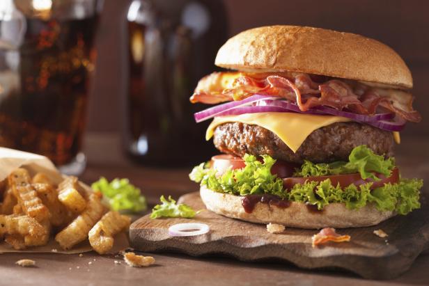 Ein Hamburger mit Speck, Käse, Tomaten und Salat, angerichtet auf einem Brett, daneben Pommes Frittes.