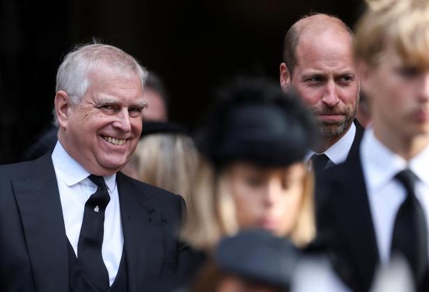 Prinz Andrew lächelt, während Prinz William ernst im Hintergrund steht.