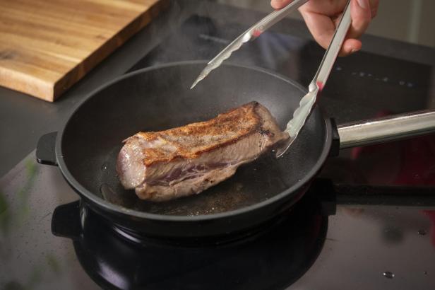 Eine Person hält Zangen über ein Steak, das in einer Pfanne brät.