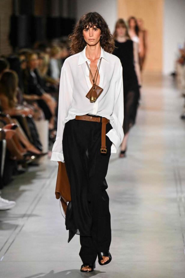 Ein Model präsentiert eine weiße Bluse und schwarze Hose auf einem Laufsteg.