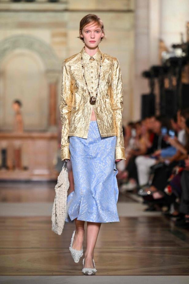 Ein Model präsentiert eine goldene Jacke und einen hellblauen Rock auf dem Laufsteg.