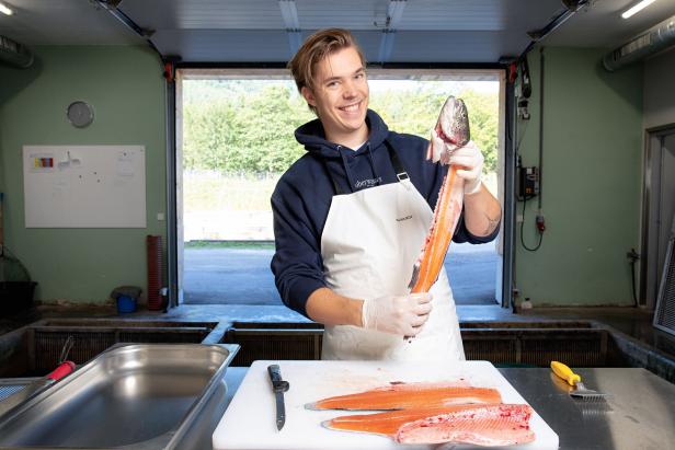 Ein lächelnder Mann mit Schürze hält einen halbierten Lachs in die Höhe.