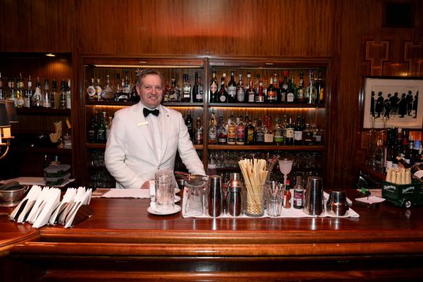 Ein Barkeeper in weißem Anzug steht hinter einer Bar mit Spirituosenflaschen.