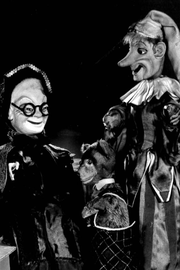 Eine Schwarzweißaufnahme von drei Marionetten, darunter eine alte Frau, ein Bär und eine Figur mit langer Nase.