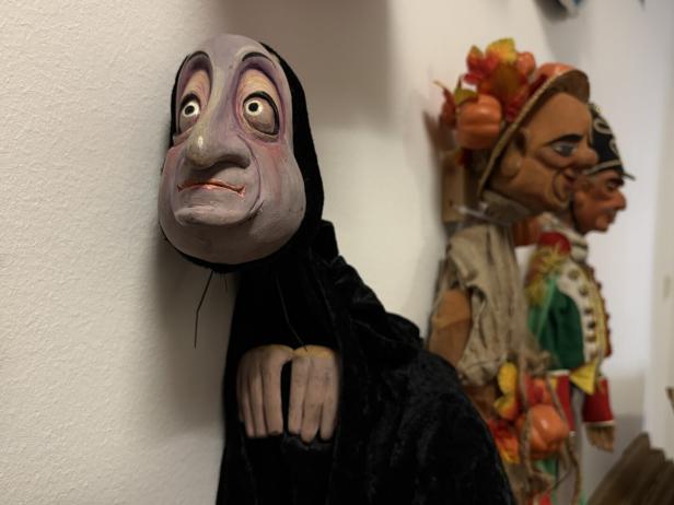 Eine Marionette mit einem ausdrucksstarken Gesicht und dunkler Kapuze hängt an einer Wand.