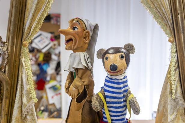 Eine Holzpuppe mit langer Nase und ein Teddybär mit blau-weiß gestreiftem Hemd stehen vor einem Vorhang.