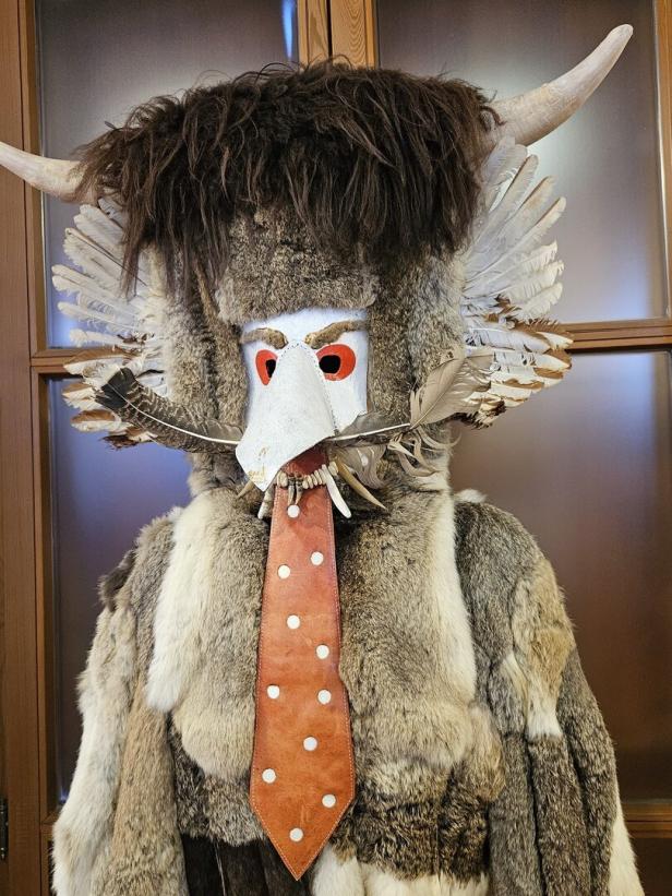 Eine aufwändige, traditionelle Maske mit Fell, Hörnern, Federn und einer Krawatte.