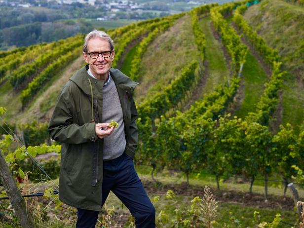 Ein Mann mit Brille steht in einem Weinberg und hält eine Traube in der Hand.