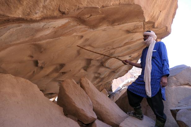 Ein Tuareg steht mit einem Stock in der Hand in einer Wüstenlandschaft.