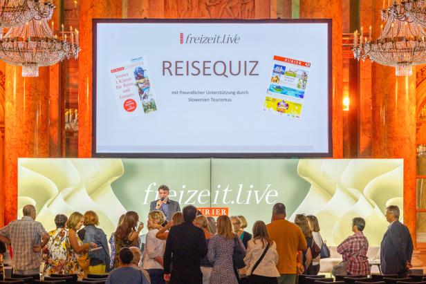 Ein Mann moderiert ein „freizeit.live“-Reisequiz vor Publikum, unterstützt von Slowenien Tourismus.