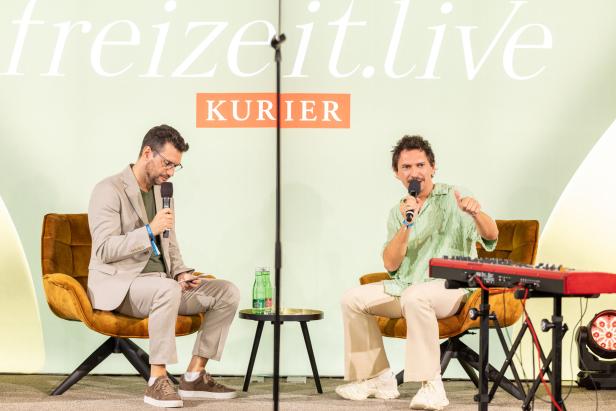 Zwei Männer sitzen auf Sesseln und sprechen bei einer Veranstaltung ins Mikrofon.