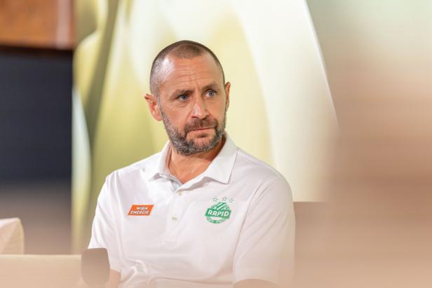 Ein Mann mit Bart und blauem Hemd mit dem Logo von Rapid Wien.