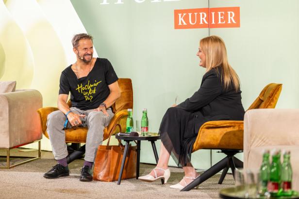 Ein Mann mit T-Shirt „Hochmair, wo bist Du?“ und eine Frau sitzen auf Sesseln und unterhalten sich.