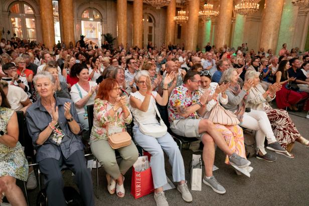 Ein großes Publikum applaudiert in einem eleganten Saal mit Säulen und Kronleuchtern.